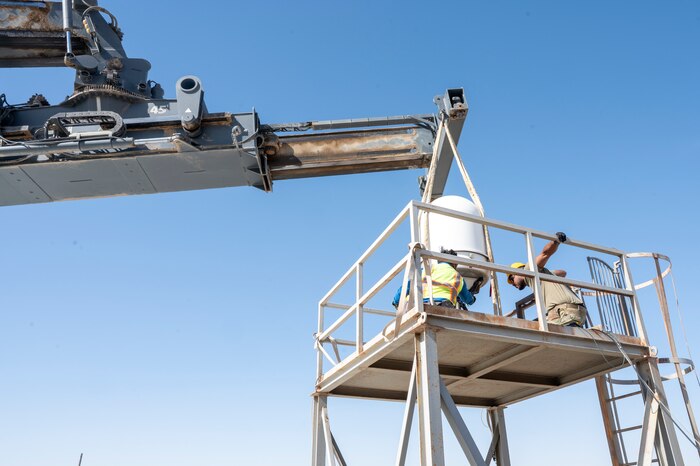 A crane left of frame lowers a PDR right of frame onto a platform where Airmen and a contractor work to install the system