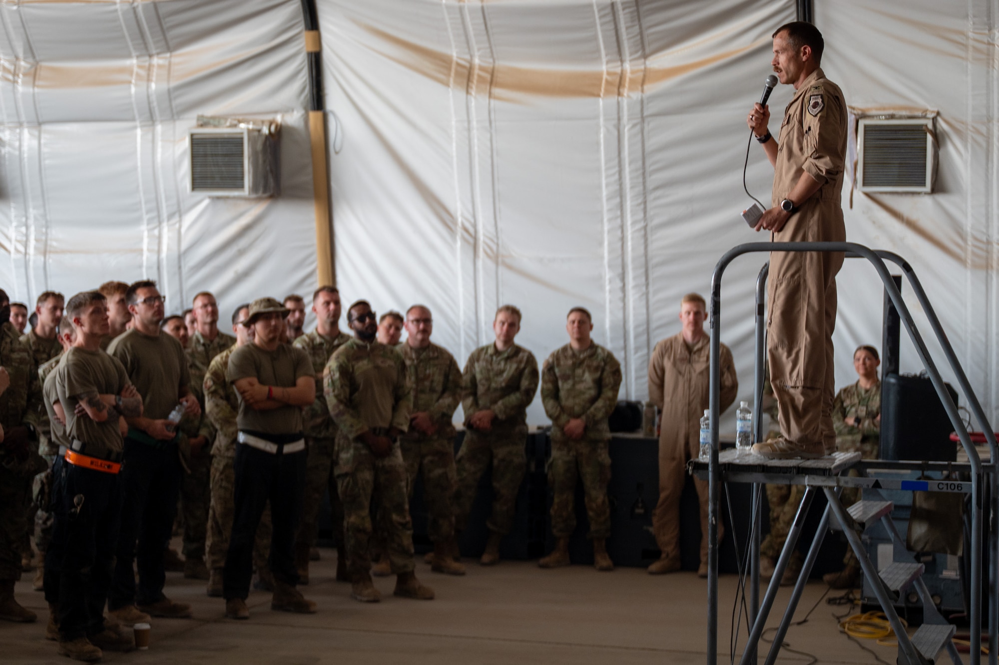 An Airman stands on a platform holding a microphone while a crowd of Airmen gather around