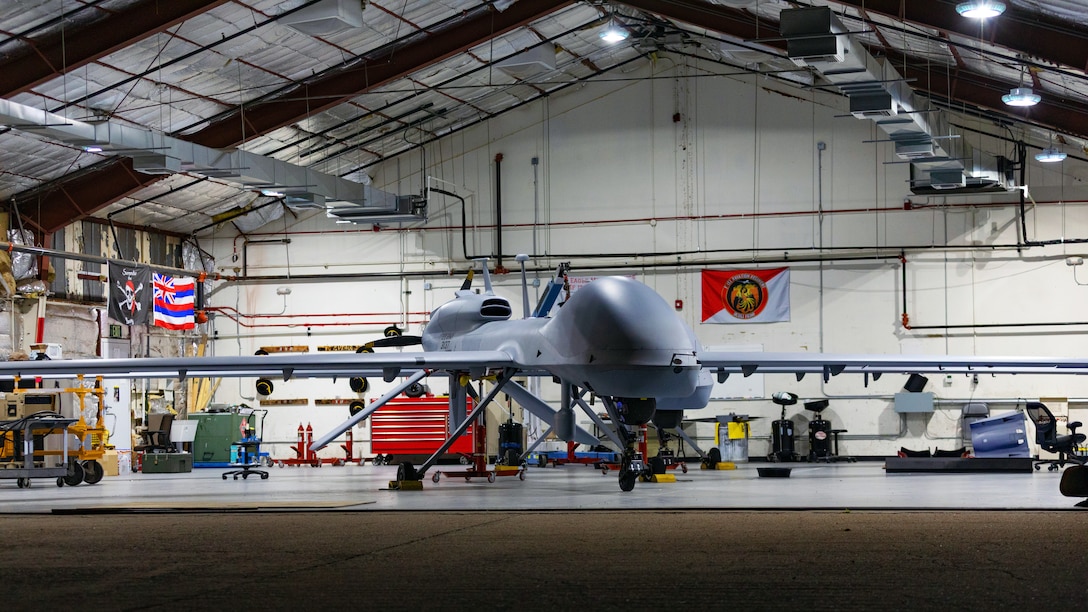 A large, gray drone aircraft is seen inside a hangar, with several flags hanging nearby and other equipment in the background.