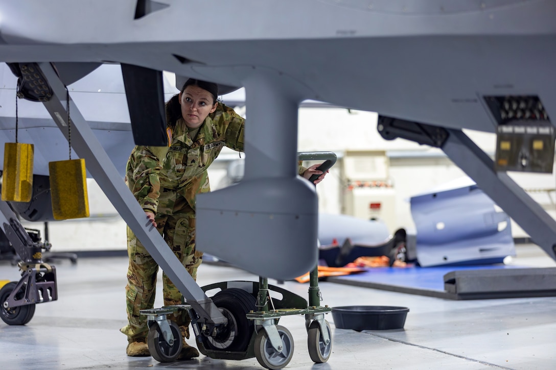 A soldier bends at the waist to look at the underside of an aircraft parked in a hangar; equipment is in the background.