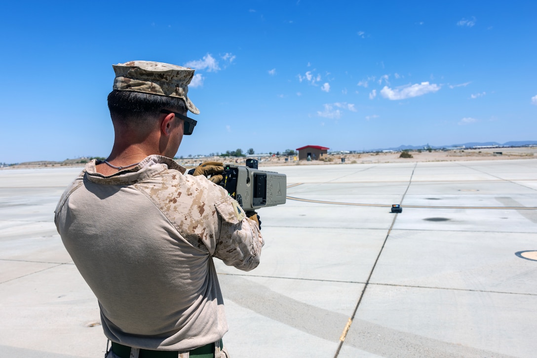 A Marine holds a device with one hand and uses the other hand to adjust a setting on the device while pointing it toward a small object on a runway on a sunny day.