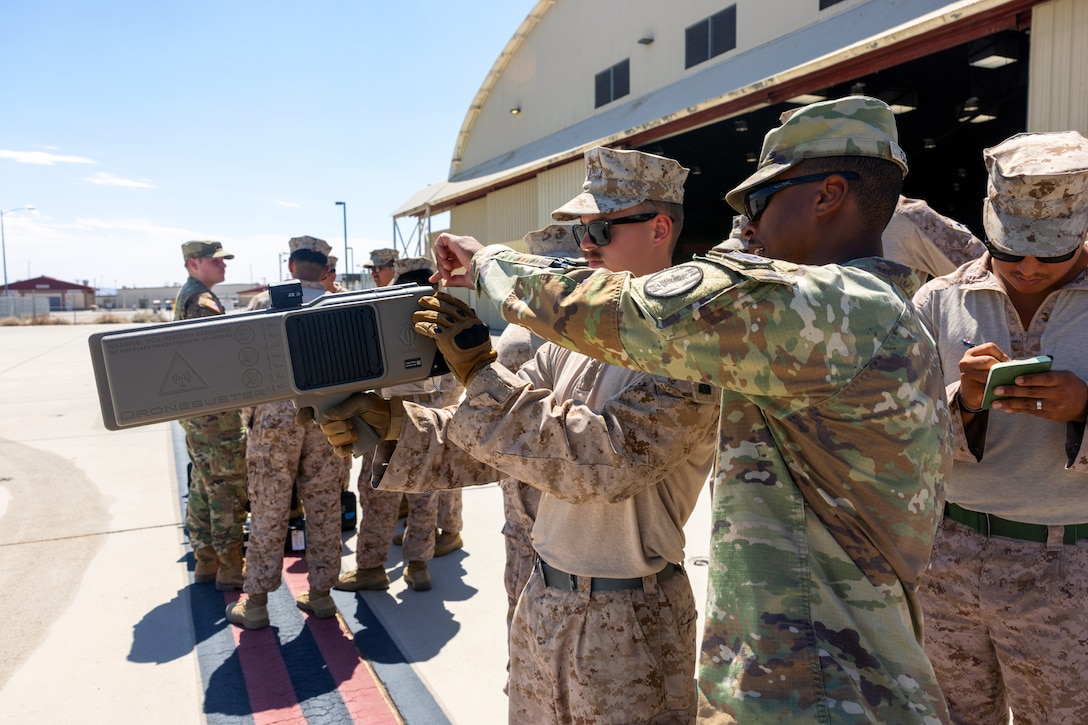 A soldier shows a Marine how to operate an anti-drone device as others stand near them at a paved area with a building in the background on a sunny day.
