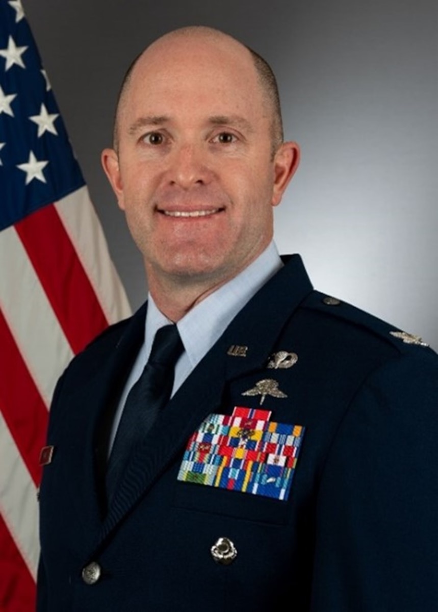 Image of man in Air Force blues uniform in front of flag.