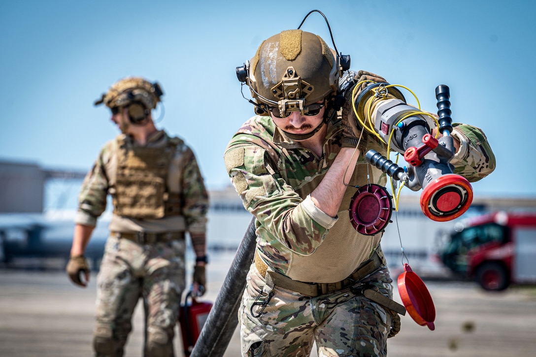 An airman drags a hose on a tarmac as a fellow airman looks to the left in the blurred background against a blue sky.