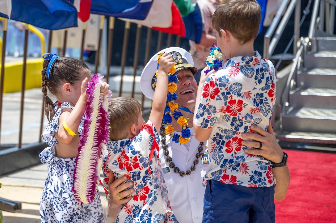 A smiling sailor kneels on a red carpet while being greeted by three children in Hawaiian gear while holding leis.