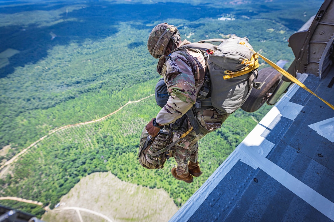 A soldier carrying gear jumps from a partially visible helicopter over a wooded area.