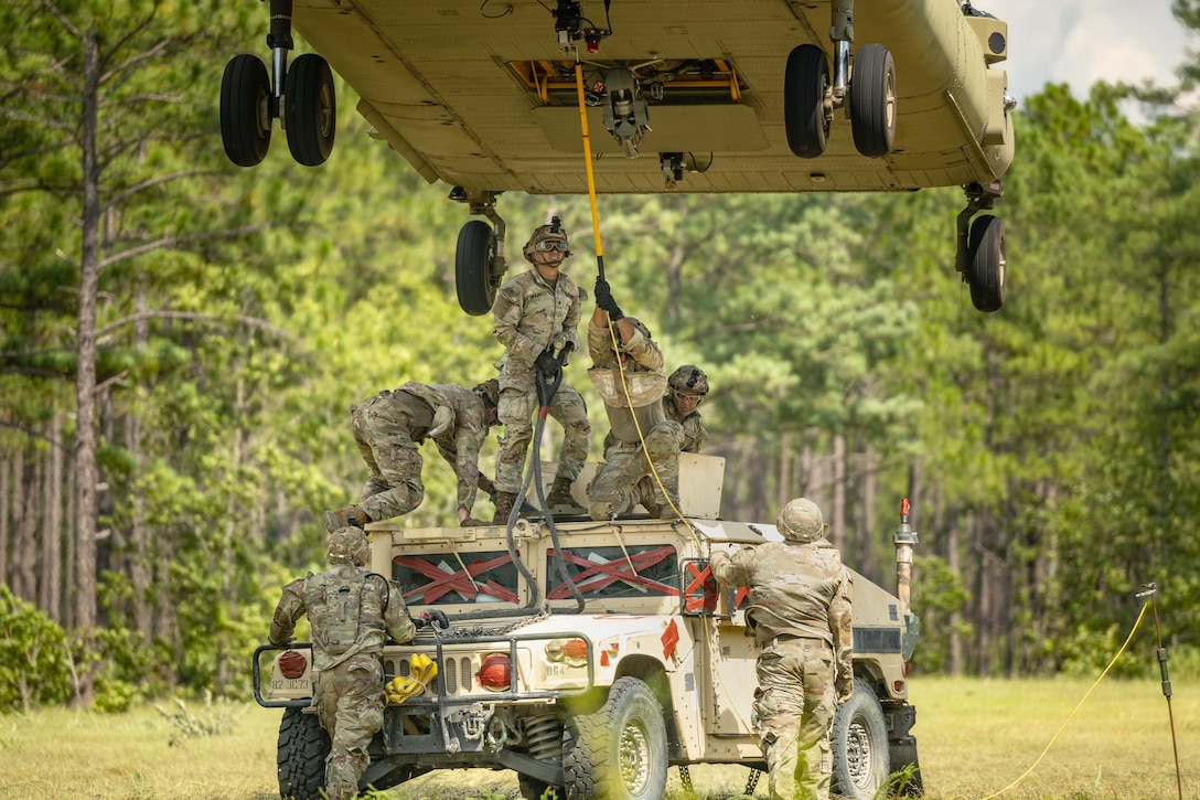Soldiers hook a vehicle up to a helicopter in a field during daylight.