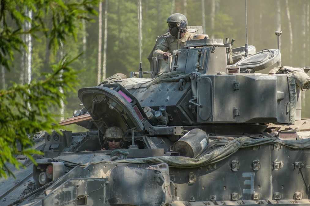 Two soldiers in helmets emerge from a military vehicle in a forest during the day.