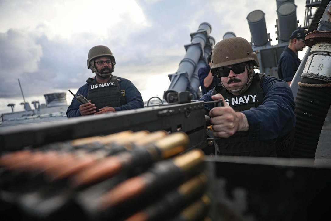 A sailor in a helmet and sunglasses aims a machine gun on a cloudy day while other soldiers stand behind.