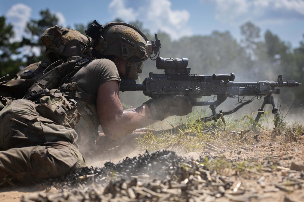 A soldier lying on the ground fires a service weapon next to a pile of bullet casings on a sunny day.