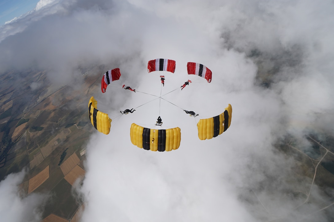 Six parachutists — three with primarily red parachutes, three with mostly yellow — connect midair, forming a circle with their canopies.