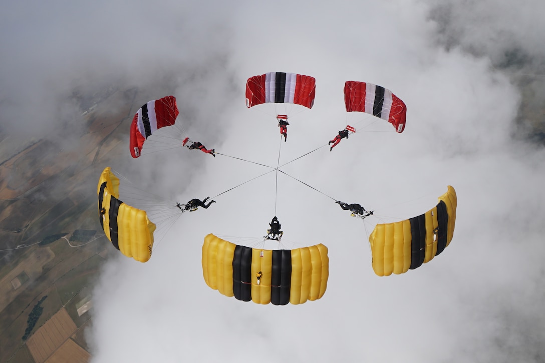 Six parachutists — three with primarily red parachutes, three with mostly yellow — connect midair, forming a circle with their canopies.