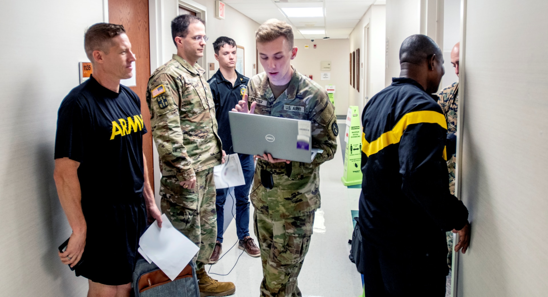 Uniformed service members stand in line with papers while medical provider holds laptop.