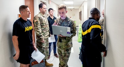 Uniformed service members stand in line with papers while medical provider holds laptop.