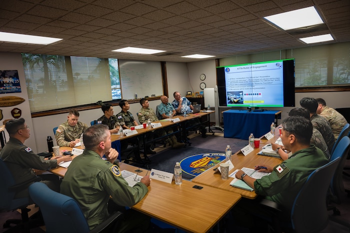 a dozen men in flight suits sit around a U-shaped conference table facing a television