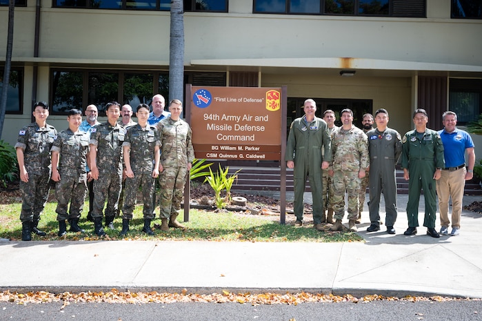 16 men, most in uniform, pose by a building sign on an army base
