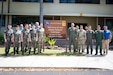 16 men, most in uniform, pose by a building sign on an army base