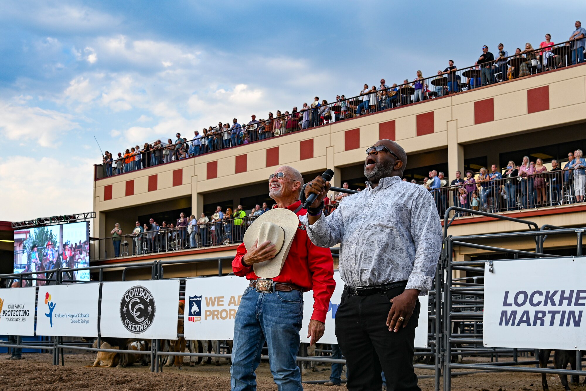A man singing in a rodeo arena as another man stands beside him with a crowd behind them.