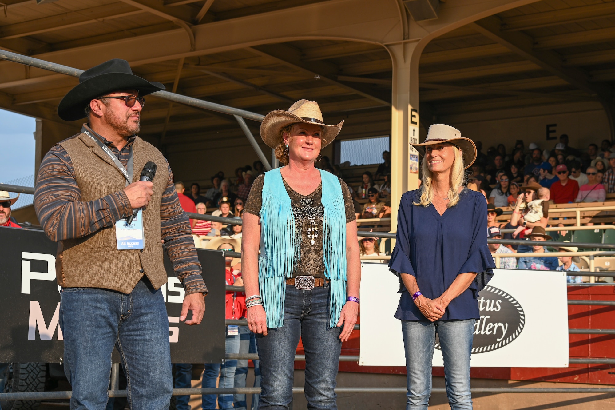 A man and two women standing in a rodeo arena with a crowd behind them in stadium seating.