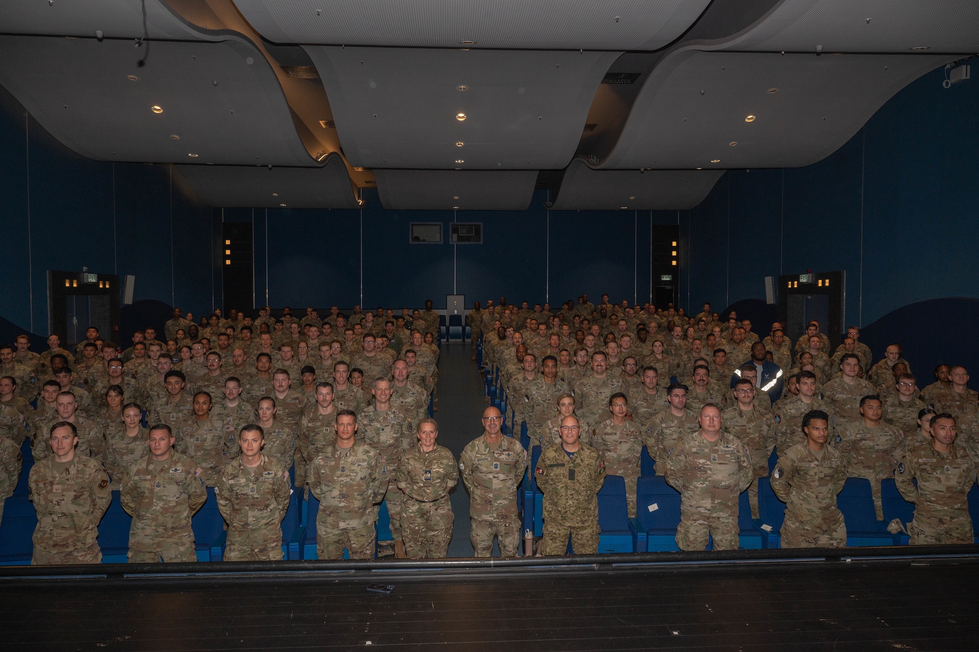 The crowd stands up from their seats, at parade rest, in an auditorium