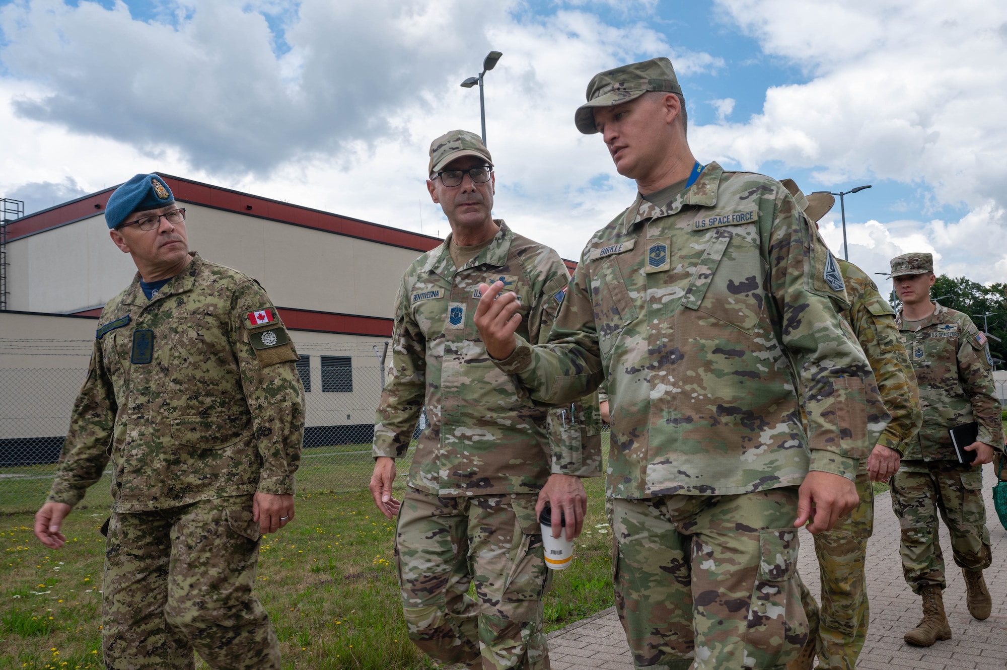 Three men at the head of a causal column of military members are walking on a sidewalk outside