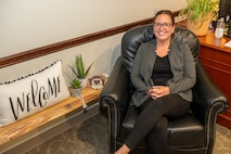 A person poses for a photo in a chair near a shelf with plants and a welcome pillow on it.