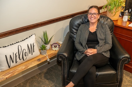 A person poses for a photo in a chair near a shelf with plants and a welcome pillow on it.