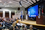 Four people stand on stage in a cross formation. A tall man in a blue Air Force uniform stands with the blue DLA Flag. To his left is a man in a dark suit, to that man's left is a man in a Army dress uniform and to his left is a woman in a Navy dress white uniform. All stand on stage ready to participate in the traditional flag pass.
