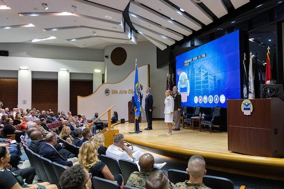Four people stand on stage in a cross formation. A tall man in a blue Air Force uniform stands with the blue DLA Flag. To his left is a man in a dark suit, to that man's left is a man in a Army dress uniform and to his left is a woman in a Navy dress white uniform. All stand on stage ready to participate in the traditional flag pass.
