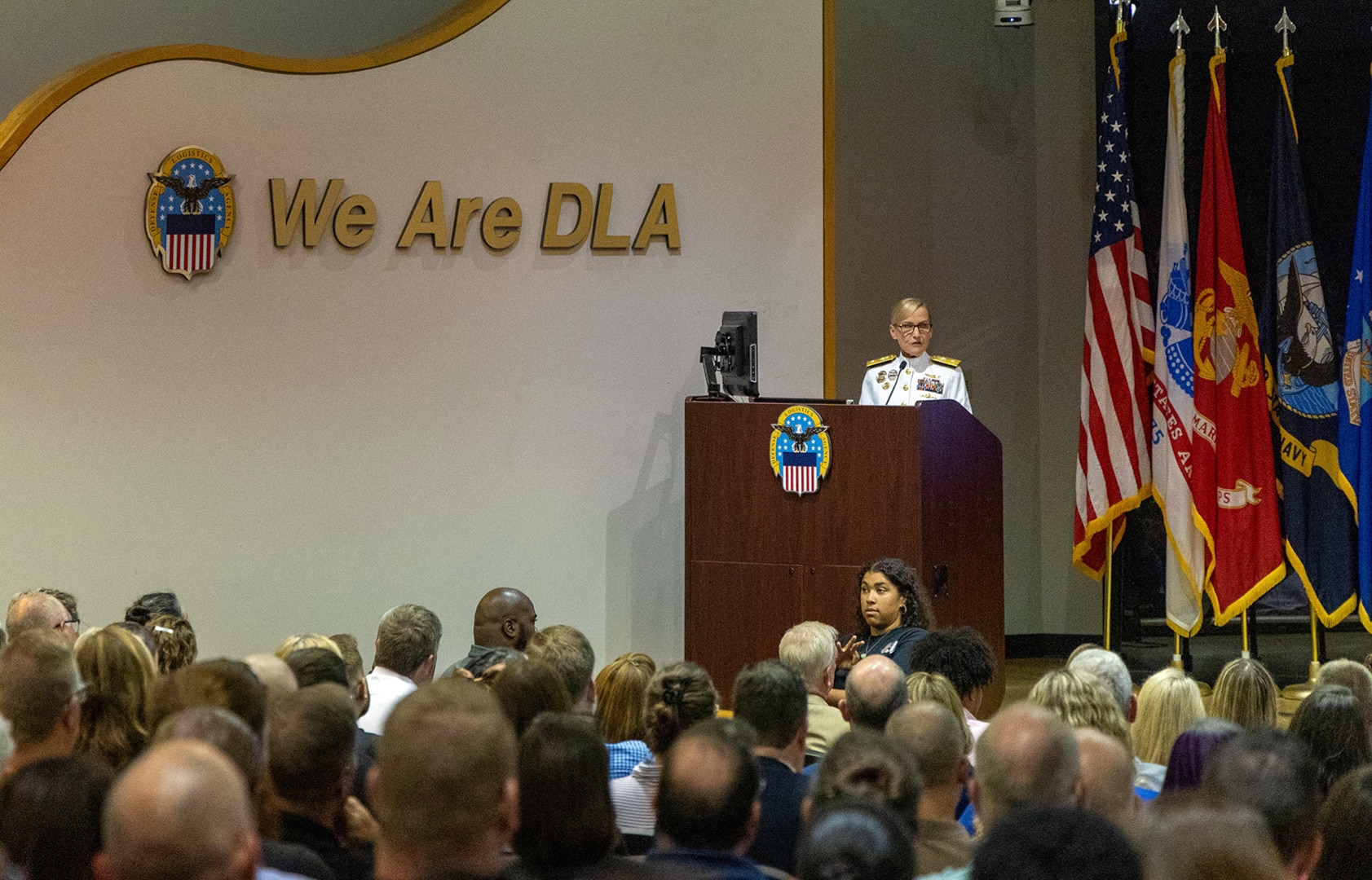 A woman in a Navy dress white uniform speaks to a full auditorium. She is at a podium on stage. There are flags behind her and to her left.