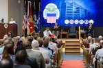 A woman in a Navy dress white uniform at a podium on a stage directs a man also in a Navy white dress uniform to hand out Ohio State Buckeye T-shirts to her family members in the front row while a man in a Army dress uniform and a man in a dark suit also on stage look on and clap.