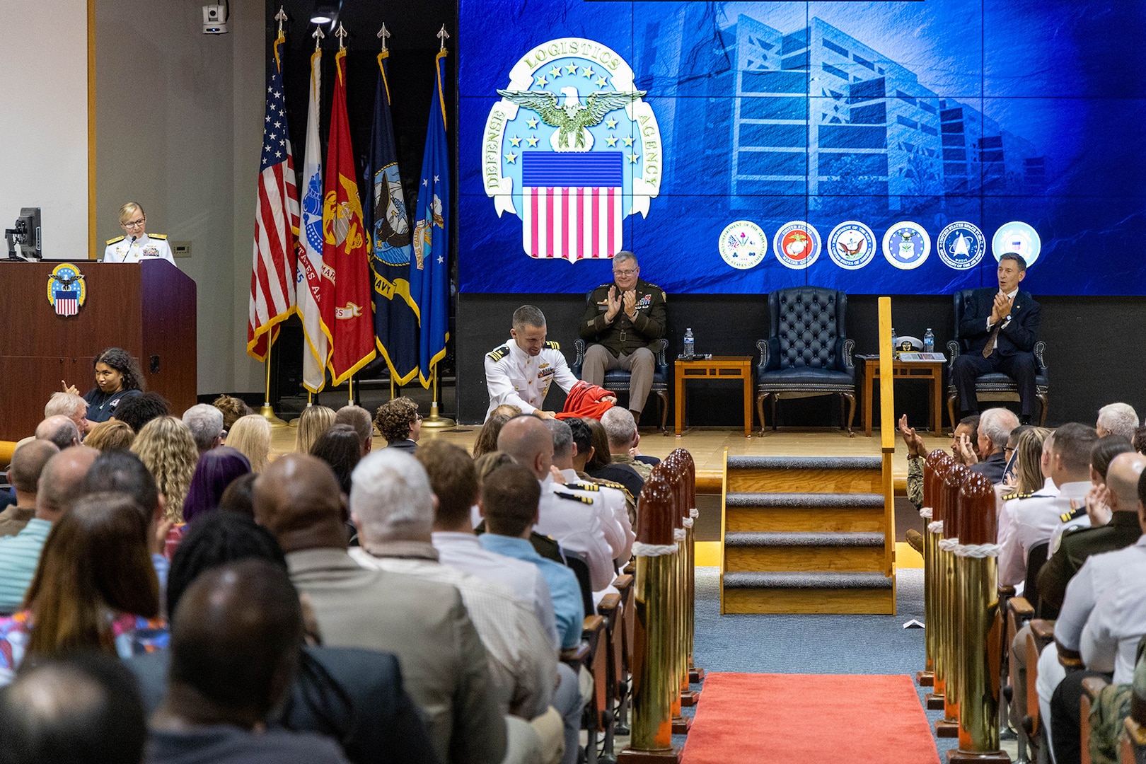 A woman in a Navy dress white uniform at a podium on a stage directs a man also in a Navy white dress uniform to hand out Ohio State Buckeye T-shirts to her family members in the front row while a man in a Army dress uniform and a man in a dark suit also on stage look on and clap.