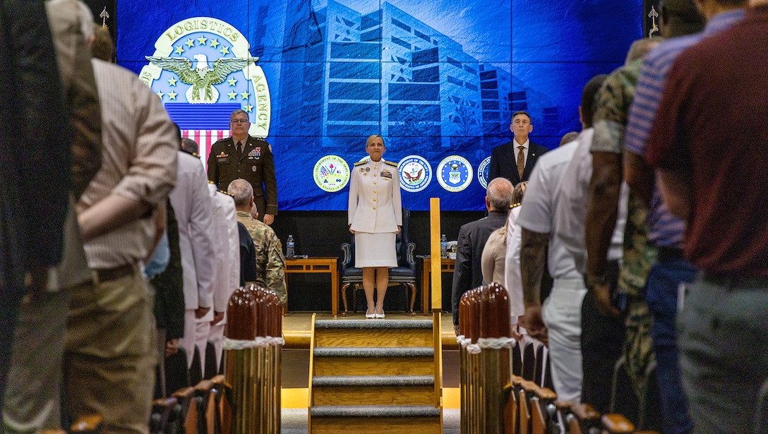 A woman dressed in Navy whites stands at attention on stage in front of a packed auditorium.