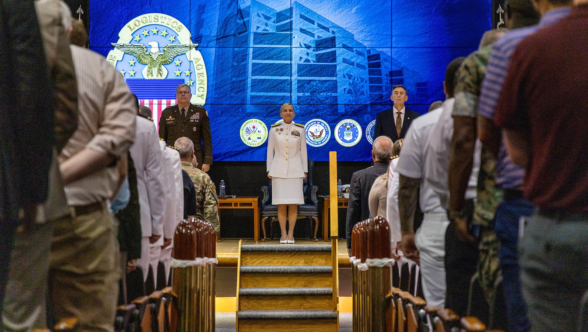 A woman dressed in Navy whites stands at attention on stage in front of a packed auditorium.
