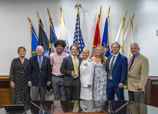A photo of a mix of people, men and women stand smiling with a woman in a Navy white dress uniform in front of a military and a U.S flag display.