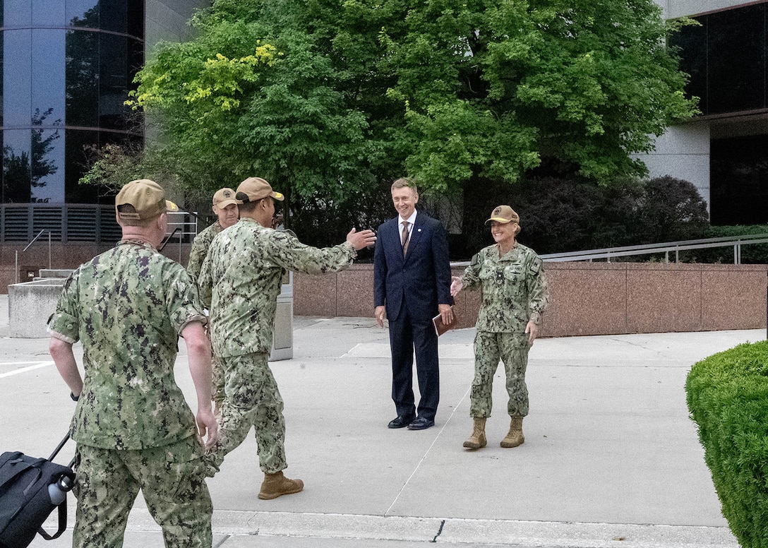 Three men in camouflaged Navy uniforms stand in front of a large gray building. One of the men reaches a woman dressed in the same type of uniform for a hug greeting while a man in a dark business suit stands next to her smiling.