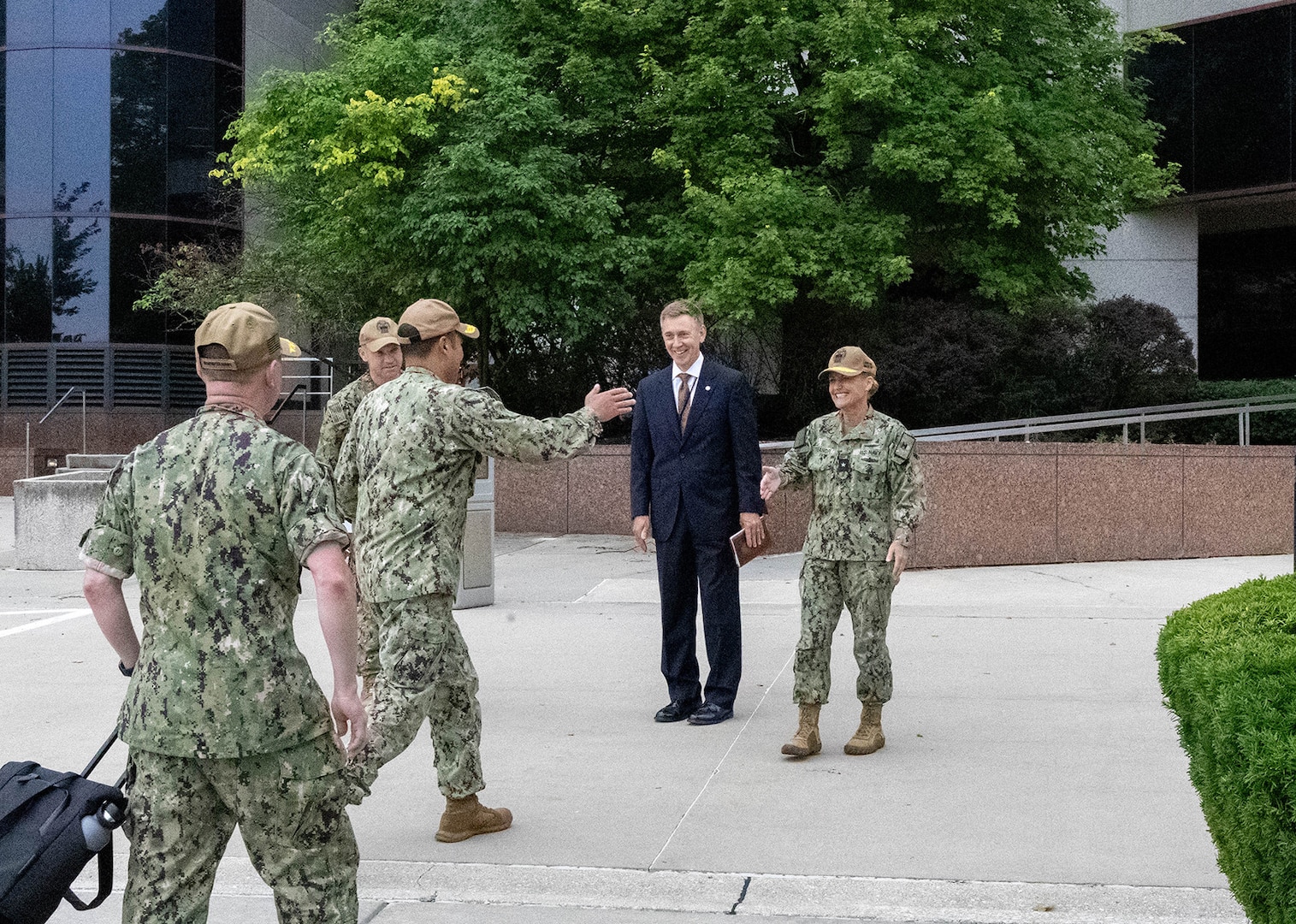 Three men in camouflaged Navy uniforms stand in front of a large gray building. One of the men reaches a woman dressed in the same type of uniform for a hug greeting while a man in a dark business suit stands next to her smiling.
