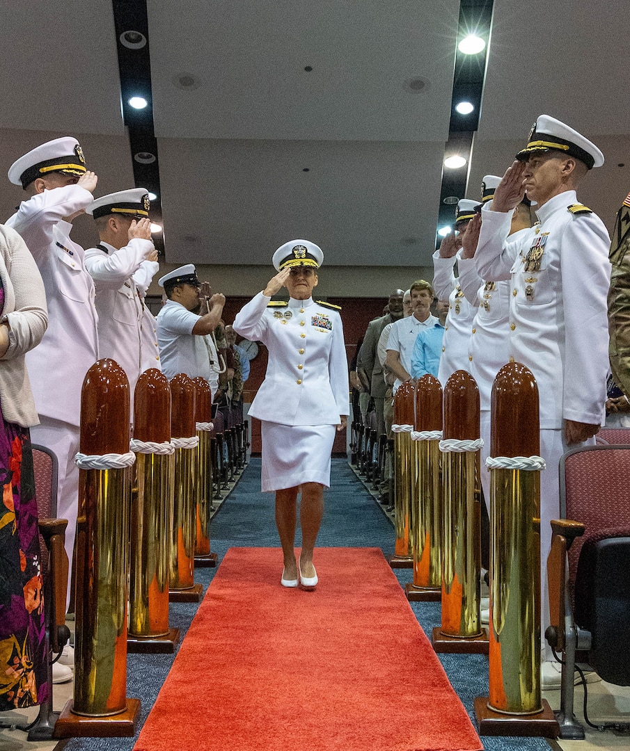 Several men dressed in Navy white dress uniforms salute while a woman dressed in Navy whites salutes as she walks by them on a red carpet lined with large replicas of shiny brass bullets in an auditorium.