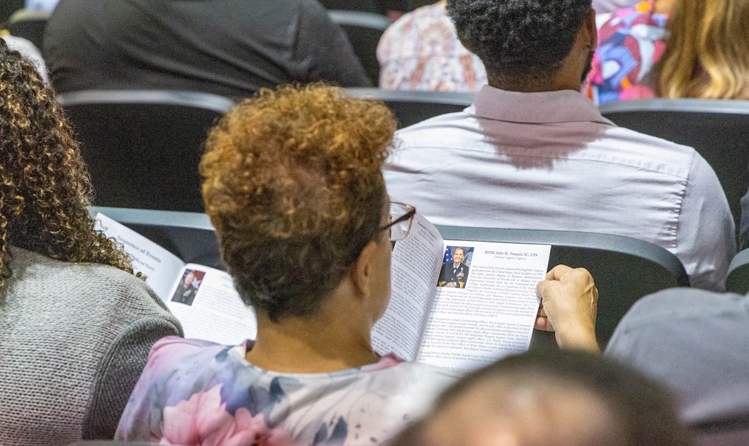 A woman with blonde curly hair looks at a program while sitting in the audience in a large auditorium.