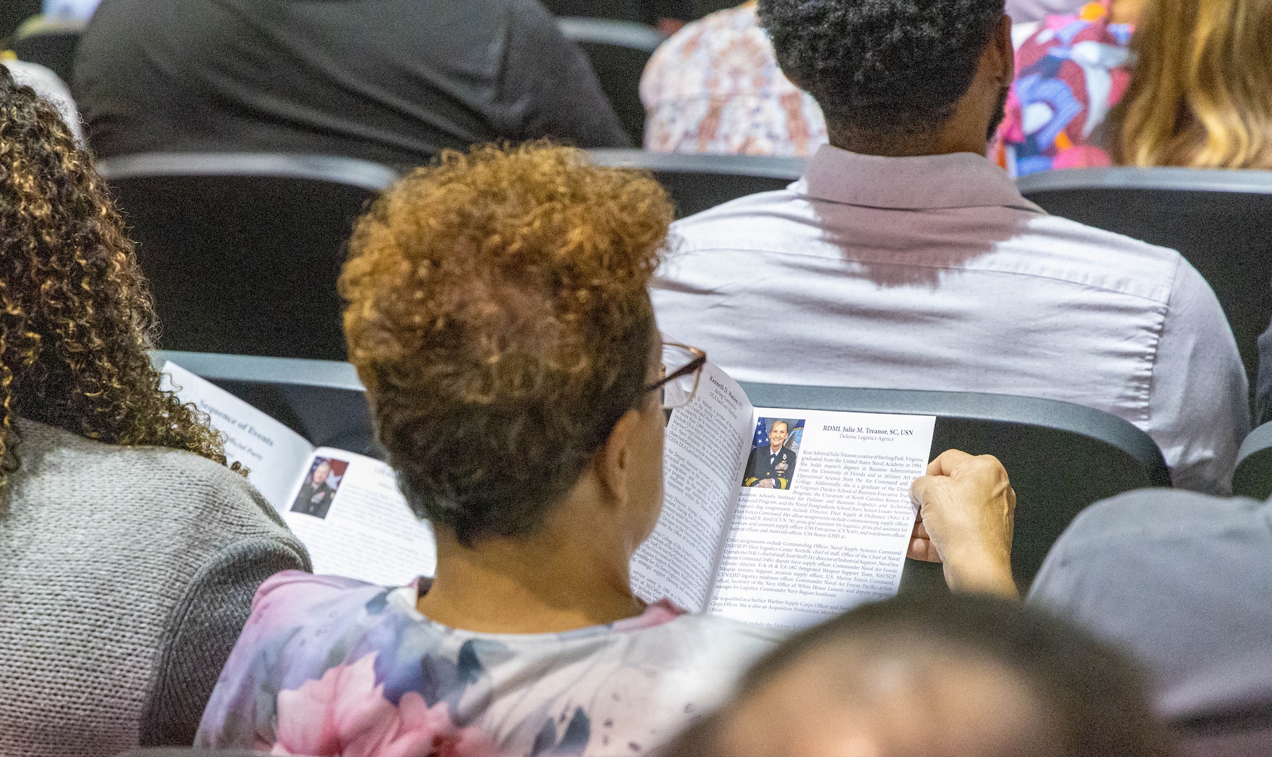 A woman with blonde curly hair looks at a program while sitting in the audience in a large auditorium.