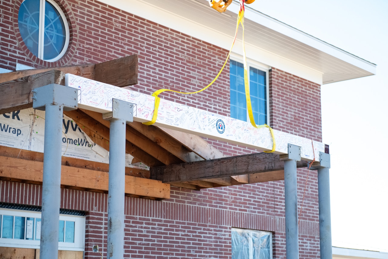 A wooden beam attached to a crane is lowered onto the top of steel poles. There is writing on the beam.