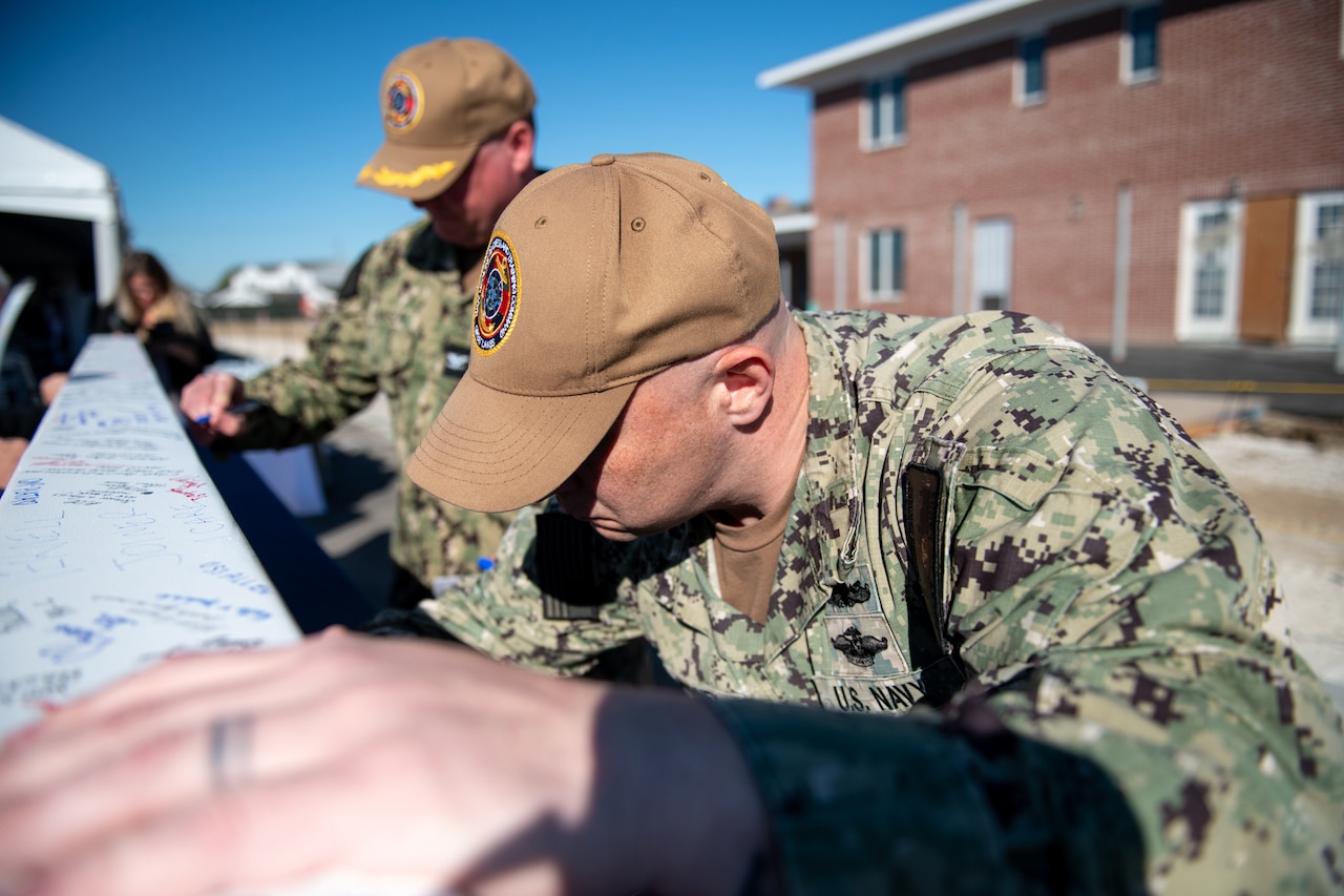 Two men in military camouflage uniforms sign a wooden beam.