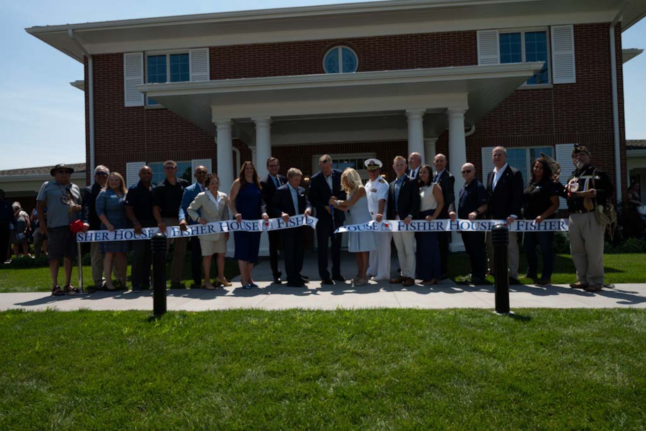 Approximately two dozen people stand behind a large ribbon as two people in the middle cut the ribbon with large scissors.