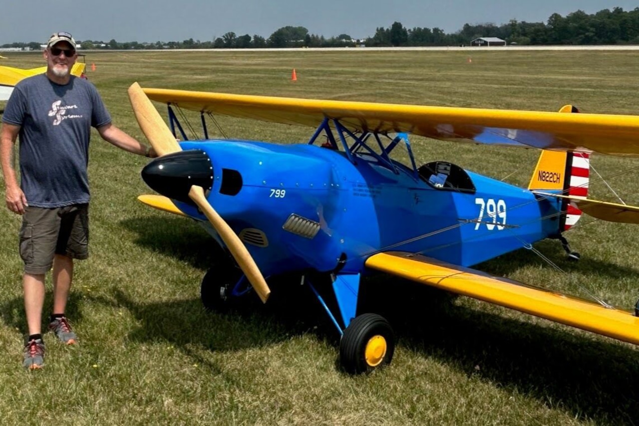 A man in shorts and a shirt stands next to a blue and yellow plane in a grass field.