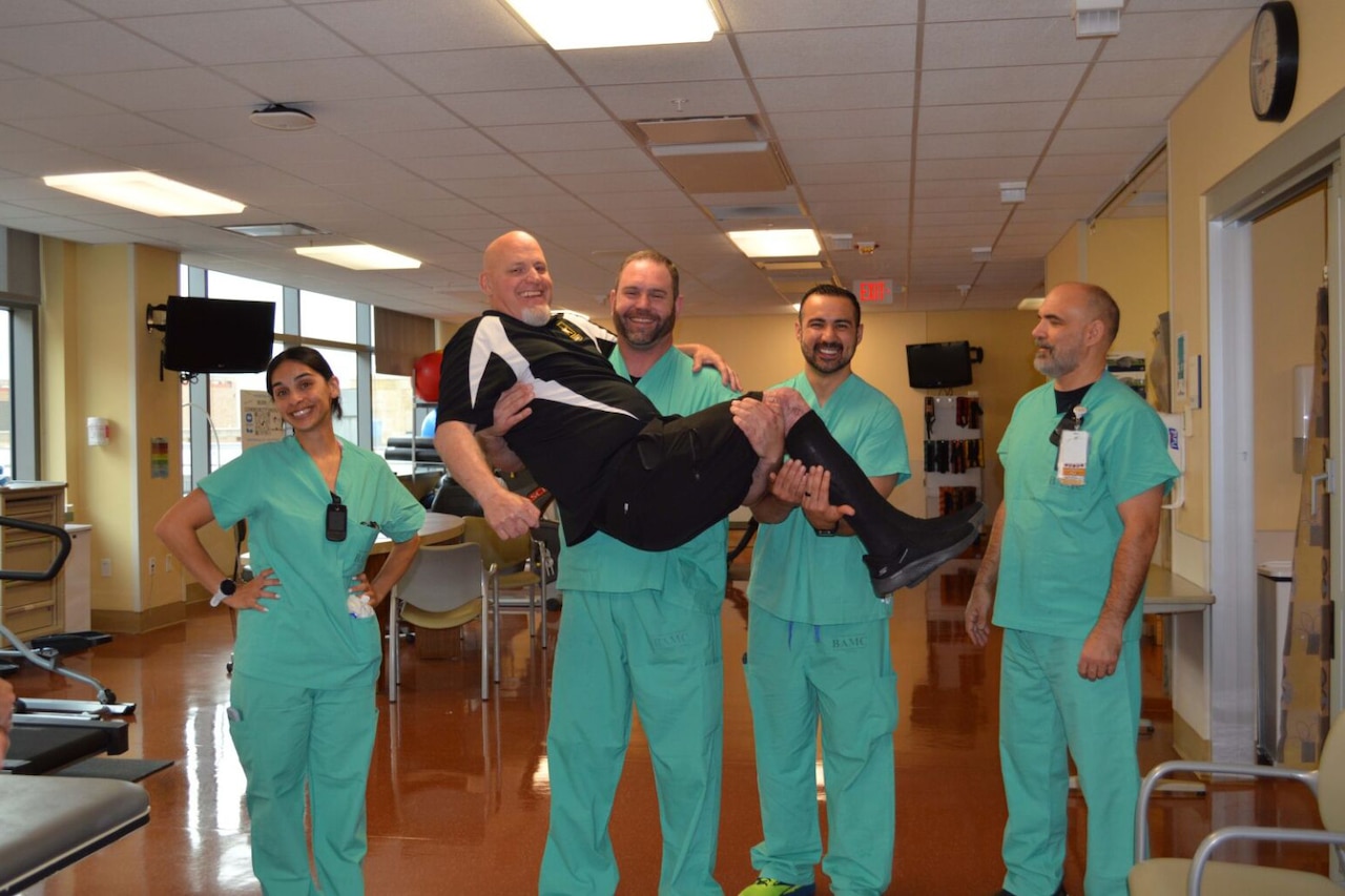 A man dressed in green scrubs lifts another man wearing black and white Army clothes while a woman and two other men, also dressed in green scrubs, smile.