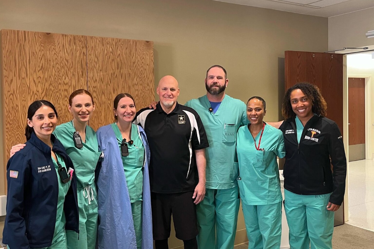 A man in black and white clothes poses with one man and seven women wearing green scrubs.