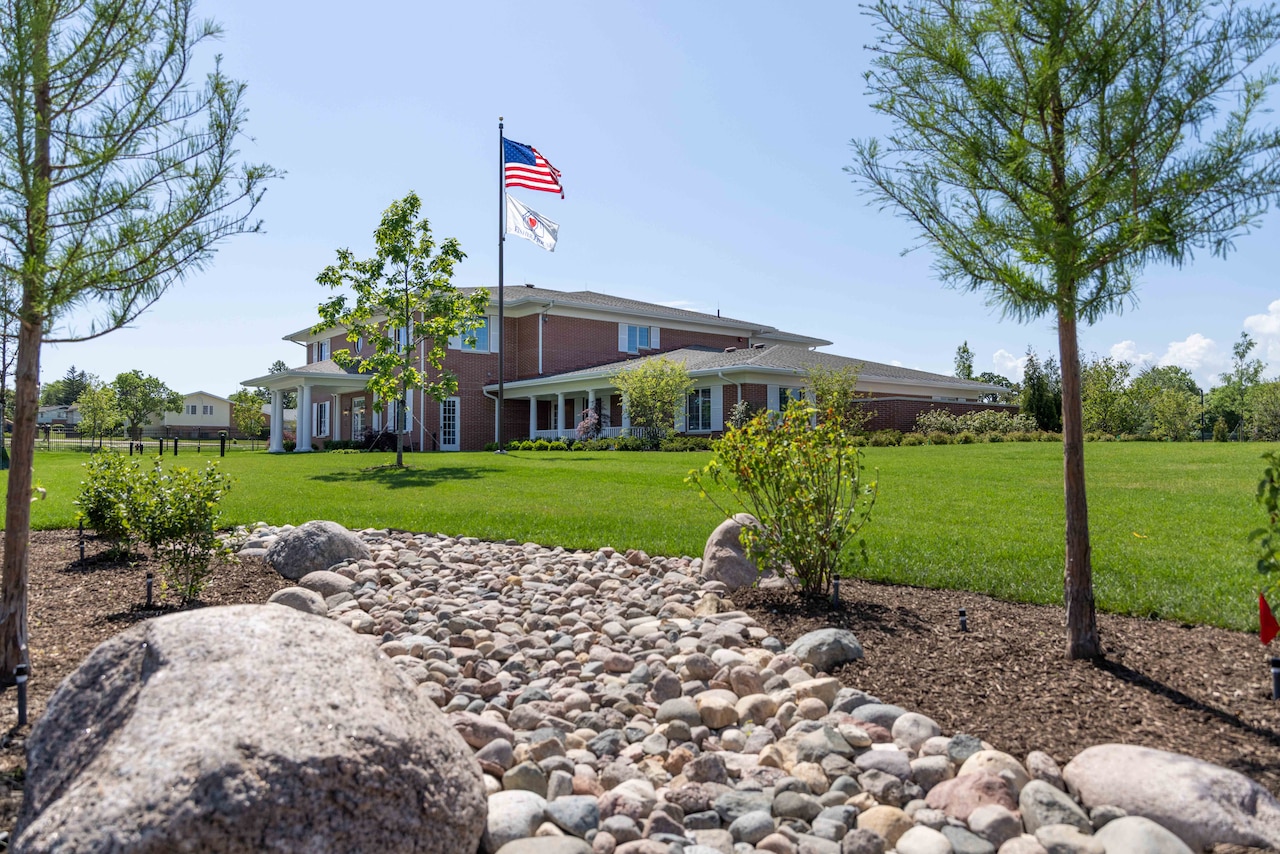 Rocks and trees dot the landscape in front of a large home with a flagpole in its yard.