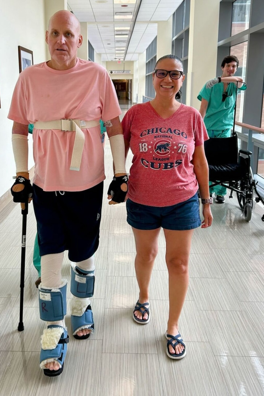 A man wrapped in bandages, support shoes and gloves walks with a cane holding hands with a man down a hospital hallway.