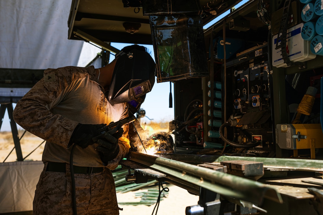 A Marine wearing face and hand protection welds a metal stake inside a tent-like structure with other equipment nearby on a sunny day outdoors.