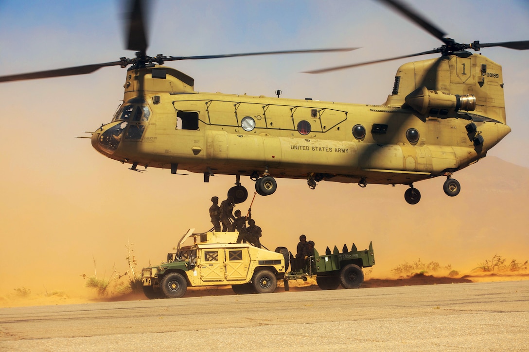 Soldiers standing atop a Humvee with a trailer attached to it work to attach a cable to the underside of a helicopter hovering above them in a desert-like area during daytime.